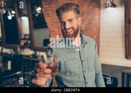 Casual men`s life. Cheers! Handsome young red bearded man in casual jeans outfit, drinking whiskey at the barber shop, enjoying and smiling Stock Photo